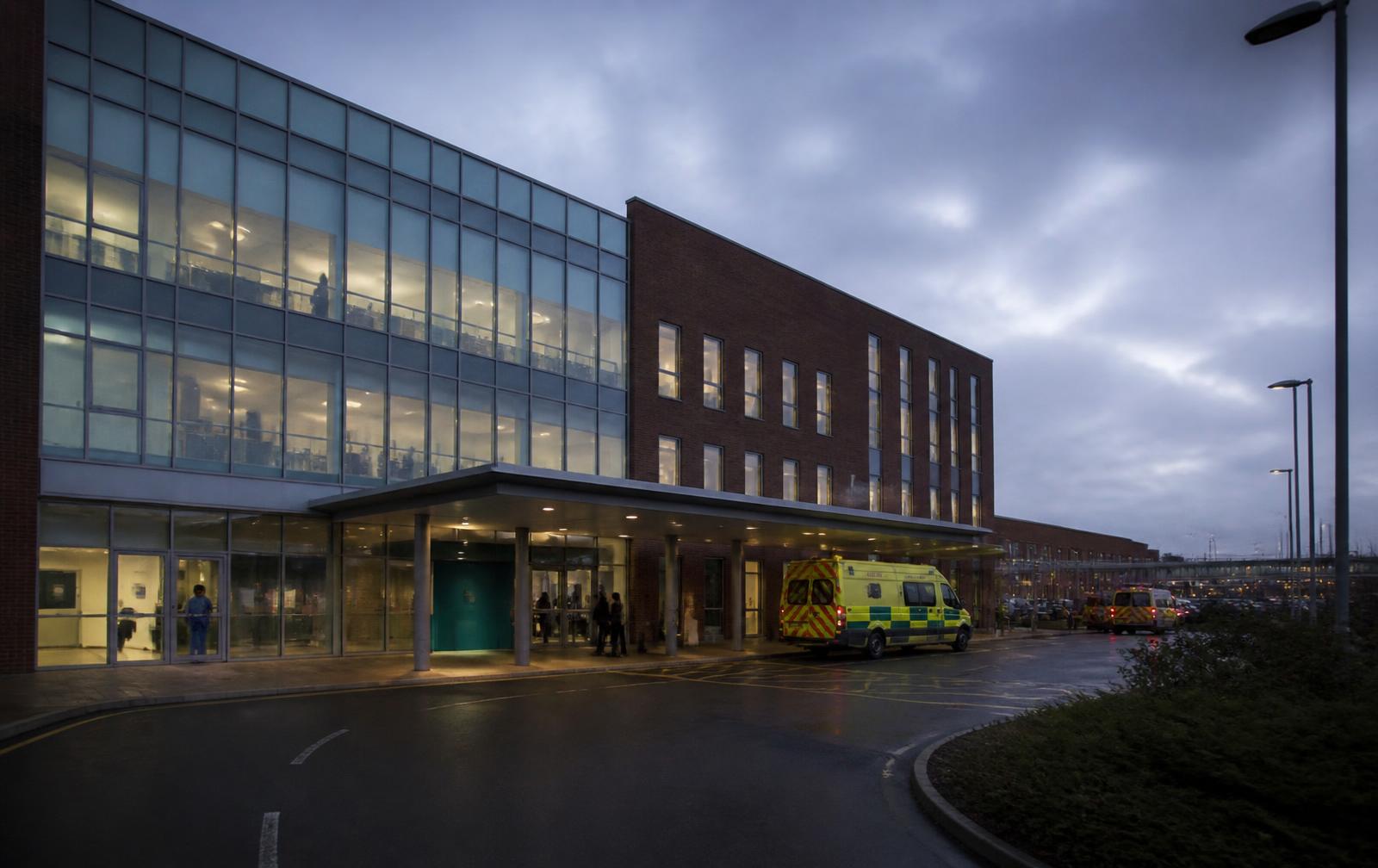 Modern UK hospital exterior at dusk with an ambulance at the entrance